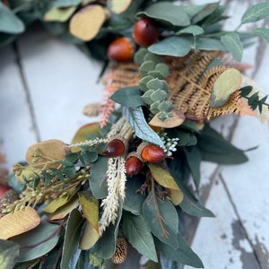 May include: A close-up of a decorative wreath featuring faux greenery, including eucalyptus leaves in shades of green and yellow. The wreath is adorned with brown acorns and dried fern accents, creating a natural, autumnal aesthetic.
