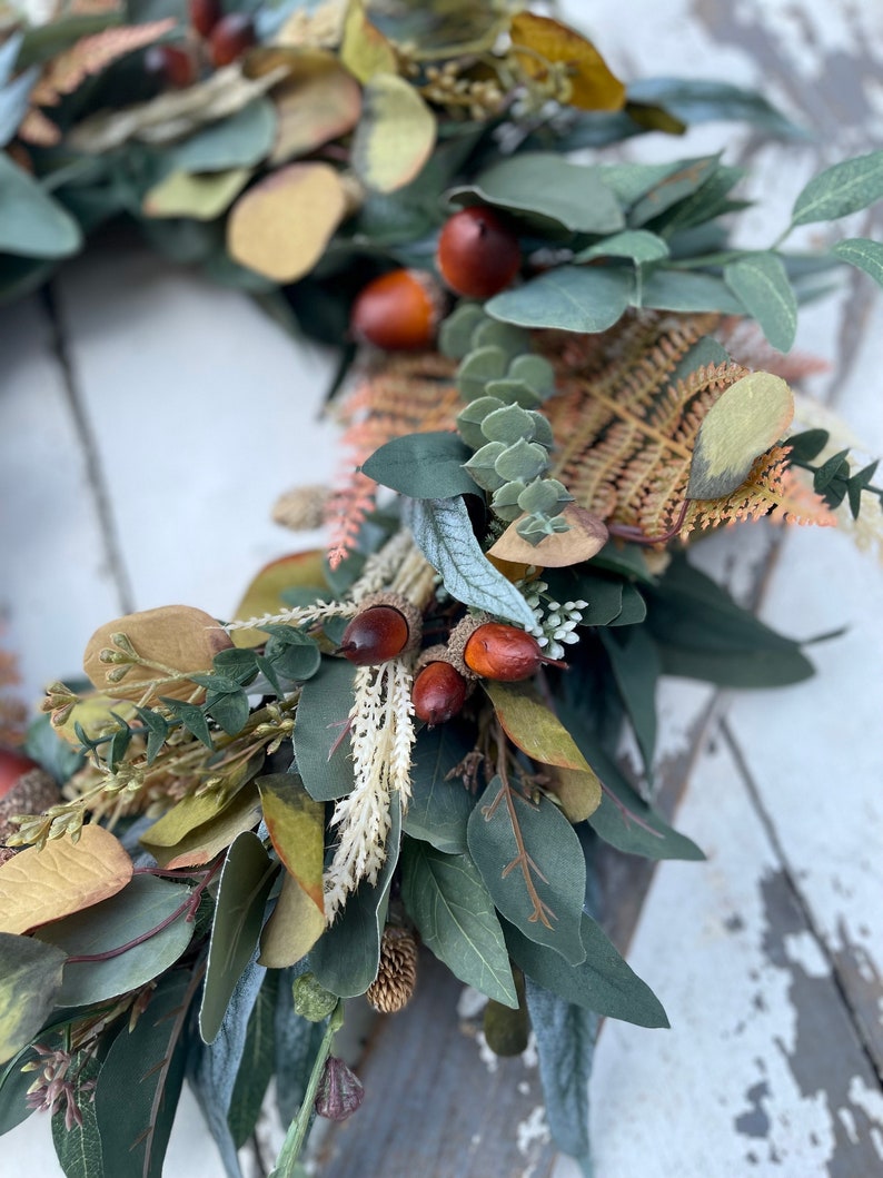 May include: A close-up of a decorative wreath featuring faux greenery, including eucalyptus leaves in shades of green and yellow. The wreath is adorned with brown acorns and dried fern accents, creating a natural, autumnal aesthetic.