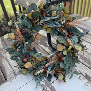 May include: A circular wreath composed of artificial foliage, including eucalyptus leaves, ferns, and small brown acorns. The wreath's colour palette is dominated by greens, with brown and tan accents, giving it a natural, autumnal appearance.