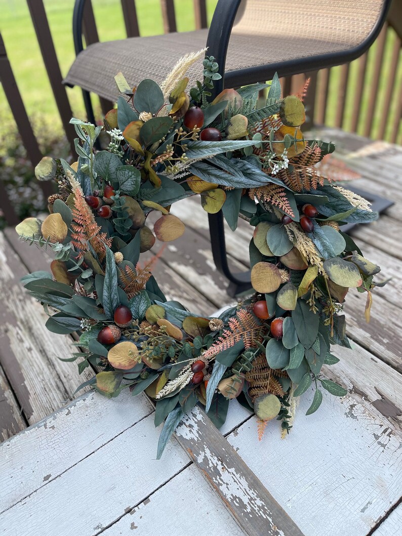 May include: A circular wreath composed of artificial foliage, including eucalyptus leaves, ferns, and small brown acorns. The wreath's colour palette is dominated by greens, with brown and tan accents, giving it a natural, autumnal appearance.