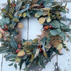 May include: A circular wreath composed of green eucalyptus leaves, brown acorns, and dried ferns. The wreath has a natural, autumnal aesthetic, with a mix of green, brown, and beige tones. The wreath is suitable for door or wall decoration.