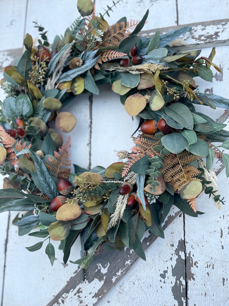 May include: A circular wreath composed of green eucalyptus leaves, brown acorns, and dried ferns. The wreath has a natural, autumnal aesthetic, with a mix of green, brown, and beige tones. The wreath is suitable for door or wall decoration.
