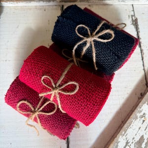 May include: Close-up of several rolls of burlap ribbon in shades of red and navy blue, tied with twine bows. The rustic texture of the burlap is visible, and the rolls are stacked on a distressed white wooden surface.