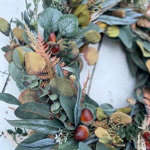 May include: A close-up of a decorative wreath with faux eucalyptus leaves in shades of green and grey, small acorns, and dried floral elements. This wreath is designed for seasonal home decor and measures approximately 30 cm in diameter.