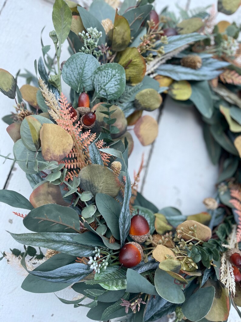 May include: A close-up of a decorative wreath with faux eucalyptus leaves in shades of green and grey, small acorns, and dried floral elements. This wreath is designed for seasonal home decor and measures approximately 30 cm in diameter.