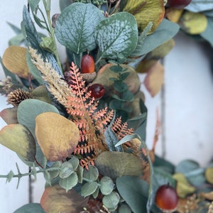 May include: A close-up of a seasonal wreath with artificial leaves and berries. The wreath features green eucalyptus leaves, brown acorns, and orange-toned sprigs. The leaves have a frosted appearance, and the colour palette is muted, with autumnal tones.