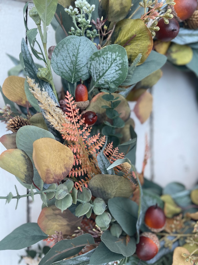 May include: A close-up of a seasonal wreath with artificial leaves and berries. The wreath features green eucalyptus leaves, brown acorns, and orange-toned sprigs. The leaves have a frosted appearance, and the colour palette is muted, with autumnal tones.