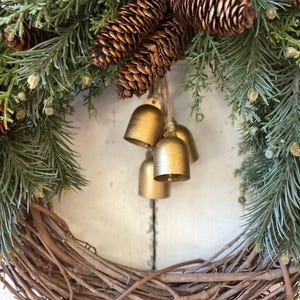May include: A wreath constructed from brown twigs, green pine needles, and pinecones, accented with bright red berries. Three small, matte gold-coloured metal bells hang from the centre of the wreath by a piece of natural twine.