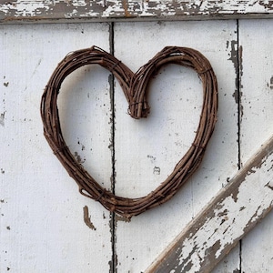 May include: A rustic, brown, heart-shaped wreath made of woven twigs. The wreath is displayed against a weathered, white-painted wooden background with visible wood grain and peeling paint, creating a charming, farmhouse aesthetic.