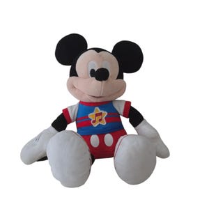 Puede incluir: Un peluche de Mickey Mouse con cara color melocot&oacute;n claro, grandes orejas negras y guantes blancos. El personaje viste una camiseta azul con rayas horizontales rojas y un parche de estrella amarilla con una nota musical roja, y pantalones rojos.