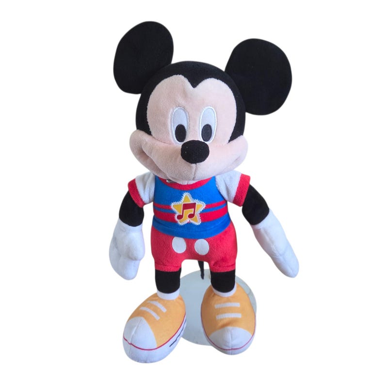 Puede incluir: Un peluche de Mickey Mouse con grandes orejas negras, cara color melocot&oacute;n y nariz negra. El personaje viste una camiseta azul con una estrella amarilla y una nota musical, pantalones cortos rojos con dos botones blancos y zapatillas amarillas.