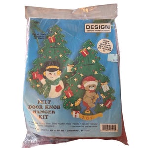 May include: A clear plastic bag containing a felt door knob hanger kit. The kit features a blue background with two green Christmas trees decorated with ornaments and a snowman and teddy bear. The text "FELT DOOR KNOB HANGER KIT" is visible.
