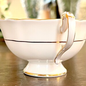 Puede incluir: Taza de t&eacute; blanca con un borde dorado y una franja negra. La taza de t&eacute; tiene un borde delicado y festoneado y un asa dorada.