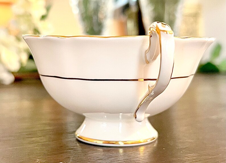 Puede incluir: Taza de t&eacute; blanca con un borde dorado y una franja negra. La taza de t&eacute; tiene un borde delicado y festoneado y un asa dorada.