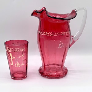 May include: A set of ruby red glass pitcher and tumbler with gold painted details. The pitcher has a clear glass handle and a ruffled rim.