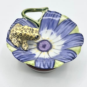 Op de afbeelding: Een keramische schotel in de vorm van een bloem met een gele kikker aan de rand. De bloem is geschilderd in tinten blauw, groen, wit en paars.
