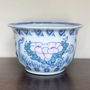 May include: A ceramic planter featuring a white base with blue and pink floral patterns. The exterior displays hand-painted peony blossoms with green leaves, surrounded by blue grass motifs and a decorative blue border along the top rim.