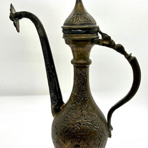 May include: An ornate brass teapot with a long, curved spout and a lid with intricate engravings. The teapot is decorated with a floral design and Arabic script.