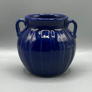 Puede incluir: Un jarrón de cerámica azul cobalto con dos asas pequeñas. El jarrón tiene un cuerpo redondeado con detalles acanalados verticales. El acabado brillante refleja la luz, resaltando las curvas y el color azul oscuro. Una pieza decorativa para la decoración del hogar.