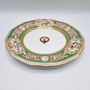 May include: A white porcelain plate with a green and pink floral design and gold accents. The center of the plate features a crest with a stag and the text "The Earl of Fife".