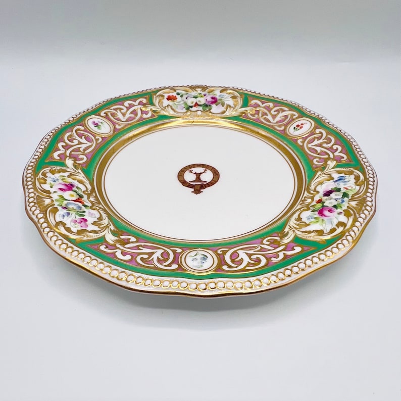 May include: A white porcelain plate with a green and pink floral design and gold accents. The center of the plate features a crest with a stag and the text "The Earl of Fife".