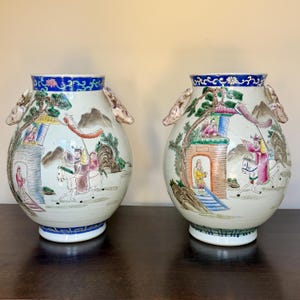 Op de afbeelding: Twee bijpassende antieke Chinese porseleinen vazen. Elke vaas heeft een bolvormig lichaam met handgeschilderde scènes van figuren te paard en in gebouwen, met berg- en boomdetails. De vazen hebben handvatten en blauw-witte randen.