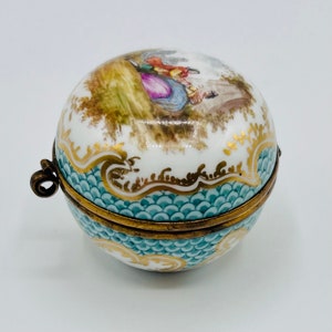 May include: A round porcelain trinket box with a hinged lid. The lid is decorated with a painted scene of people in a garden. The box is decorated with a blue and gold pattern.