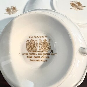 Puede incluir: Una taza de t&eacute; y un platillo de porcelana blanca con ribetes dorados y las palabras "PARAGON BY APPOINTMENT TO H.THE QUEEN & H.M QUEEN MARY FINE BONE CHINA ENGLAND REGD."