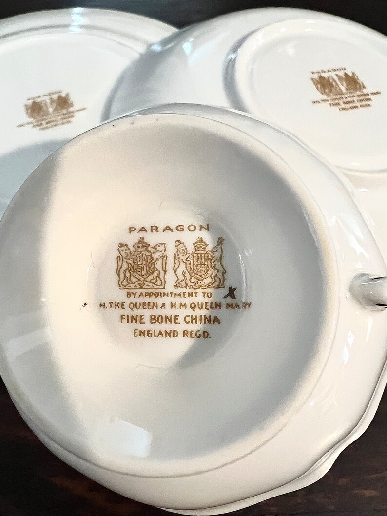 Puede incluir: Una taza de t&eacute; y un platillo de porcelana blanca con ribetes dorados y las palabras "PARAGON BY APPOINTMENT TO H.THE QUEEN & H.M QUEEN MARY FINE BONE CHINA ENGLAND REGD."