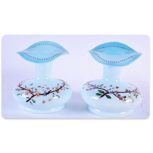 Puede incluir: Dos jarrones de cristal azul claro a juego con una forma única. Cada jarrón tiene una base redonda, un cuello delgado y una parte superior ensanchada en forma de hoja. Las bases están decoradas con diseños florales pintados a mano en tonos marrón, verde y naranja.