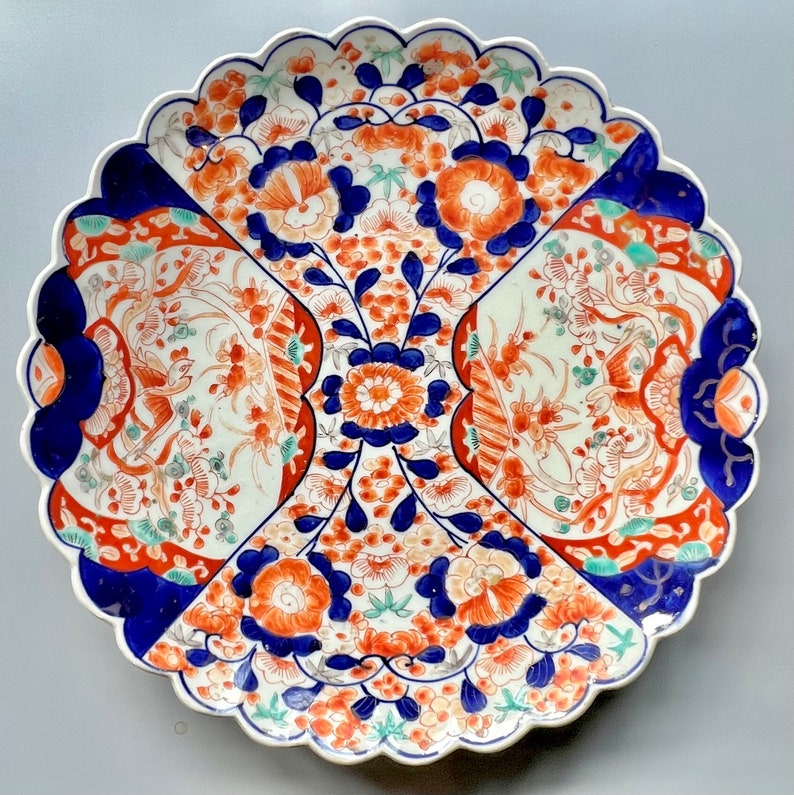 K&ouml;nnte beinhalten: Ein gro&szlig;er, wei&szlig;er Teller aus Keramik mit einem gewellten Rand. Der Teller ist mit einem farbenfrohen Blumenmuster in Blau-, Rot- und Oranget&ouml;nen verziert. Das Design zeichnet sich durch komplizierte Details und eine traditionelle japanische &Auml;sthetik aus.