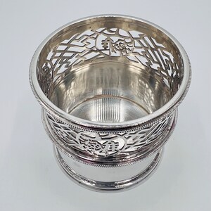 May include: Silver-plated, openwork, circular wine glass holder with a decorative design. The holder has a round base and a raised rim.