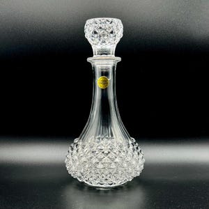 May include: A clear glass decanter with a diamond-cut pattern on the base and stopper. The decanter has a slender neck and a round, bulbous base. A small, round, gold-coloured sticker is affixed to the neck. The stopper is diamond-cut and fits snugly.
