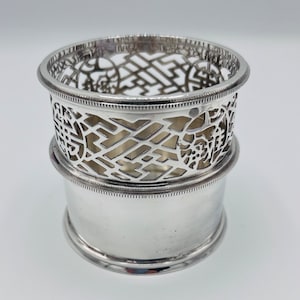 May include: Silver, decorative, openwork, cylindrical, bottle coaster with a geometric design. The coaster is made of two pieces, one that sits on top of the other.