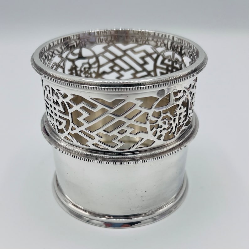 May include: Silver, decorative, openwork, cylindrical, bottle coaster with a geometric design. The coaster is made of two pieces, one that sits on top of the other.