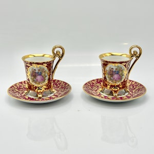 Puede incluir: Dos tazas de espresso y platillos antiguos. Las tazas y los platillos son de color burdeos con detalles dorados y un diseño detallado. Cada taza presenta un asa dorada y una pequeña pintura de una pareja. Los platillos tienen un patrón a juego.