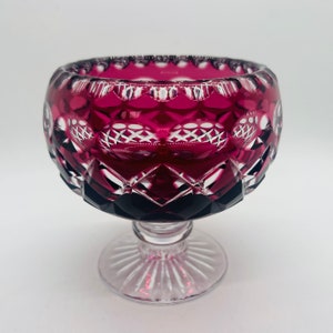 May include: A deep red crystal bowl with a cut glass design sits on a clear pedestal base. The bowl has a wide, round shape and intricate cut glass patterns.