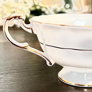 Puede incluir: Una taza de t&eacute; de porcelana blanca con un borde dorado y una banda dorada en el medio. La taza tiene un asa delicada.