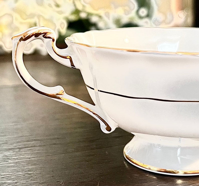 Puede incluir: Una taza de t&eacute; de porcelana blanca con un borde dorado y una banda dorada en el medio. La taza tiene un asa delicada.