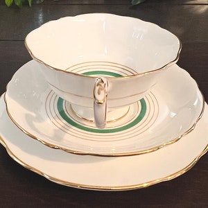 Puede incluir: Un juego de taza y platillo de t&eacute; blanco con un dise&ntilde;o de banda verde y dorada. El juego tiene un borde delicado y festoneado.