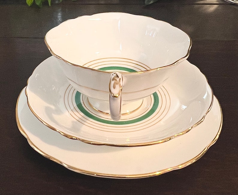 Puede incluir: Un juego de taza y platillo de t&eacute; blanco con un dise&ntilde;o de banda verde y dorada. El juego tiene un borde delicado y festoneado.