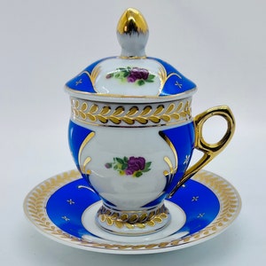 Puede incluir: Un juego de taza y platillo de porcelana con tapa. La taza, el platillo y la tapa presentan una base blanca con detalles azules y dorados. El diseño incluye patrones florales de rosas moradas. El asa y el pomo de la tapa son dorados.