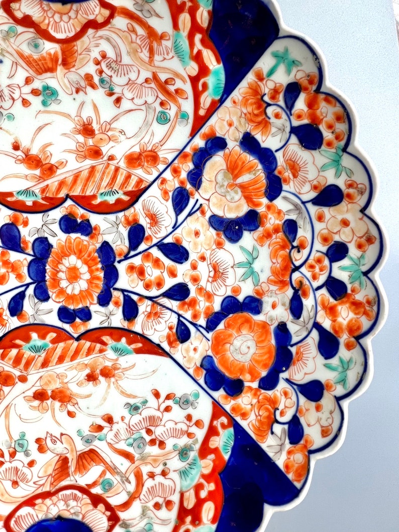 K&ouml;nnte beinhalten: Ein gro&szlig;er, wei&szlig;er Teller aus Keramik mit einem gewellten Rand. Der Teller ist mit einem farbenfrohen Blumenmuster in Blau-, Rot-, Orange- und Gr&uuml;nt&ouml;nen verziert. Das Muster umfasst Blumen, Bl&auml;tter und V&ouml;gel.