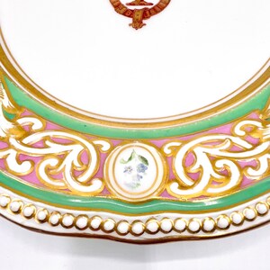 May include: A white porcelain plate with a green and pink border. The border is decorated with gold leaf and a floral design. The plate has a scalloped edge.