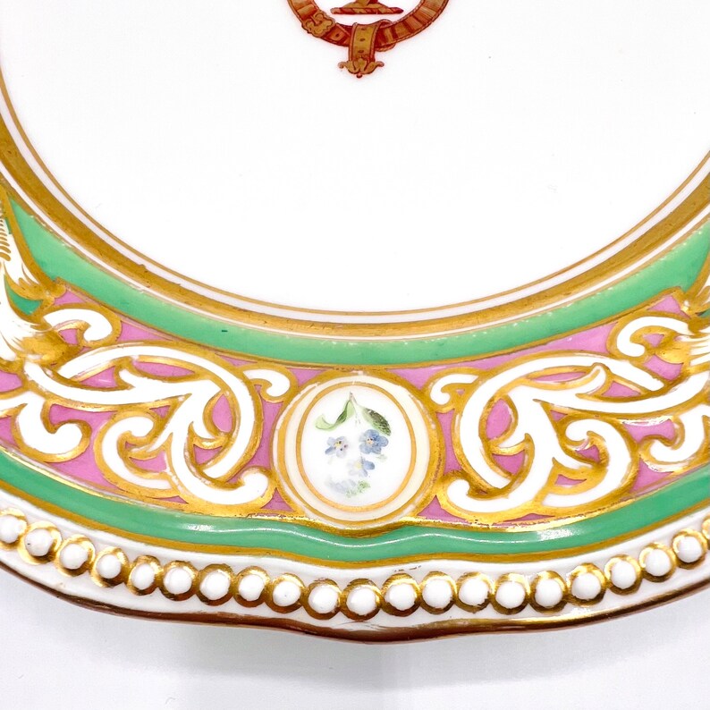 May include: A white porcelain plate with a green and pink border. The border is decorated with gold leaf and a floral design. The plate has a scalloped edge.