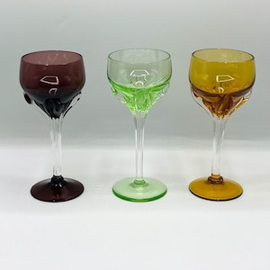 May include: Three colorful wine glasses with clear stems and bases. The glasses have bowls in shades of purple, green, and amber. The stems have a decorative design, and the bases are circular.