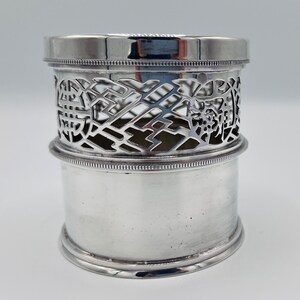 May include: Silver, openwork, cylindrical, bottle coaster with a decorative design. The coaster has a smooth, polished finish.