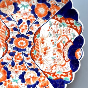 K&ouml;nnte beinhalten: Ein gro&szlig;er, wei&szlig;er Teller aus Keramik mit einem gewellten Rand. Der Teller ist mit einem farbenfrohen Blumenmuster in Blau-, Rot- und Oranget&ouml;nen verziert. Die Blumen sind stilisiert und kunstvoll, und das Design erinnert an traditionelle japanische Imari-Porzellan.