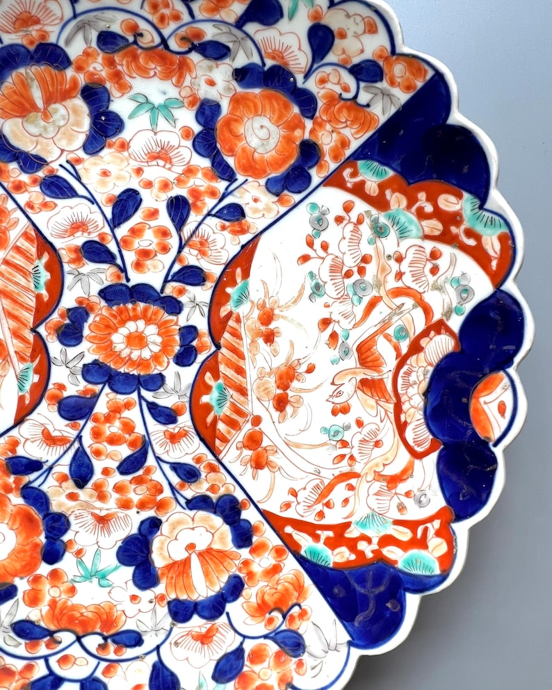 K&ouml;nnte beinhalten: Ein gro&szlig;er, wei&szlig;er Teller aus Keramik mit einem gewellten Rand. Der Teller ist mit einem farbenfrohen Blumenmuster in Blau-, Rot- und Oranget&ouml;nen verziert. Die Blumen sind stilisiert und kunstvoll, und das Design erinnert an traditionelle japanische Imari-Porzellan.