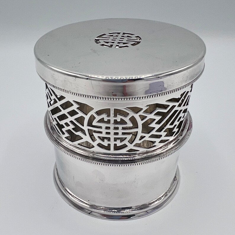 May include: Silver round container with a decorative openwork design. The container has a lid with a raised circular design.